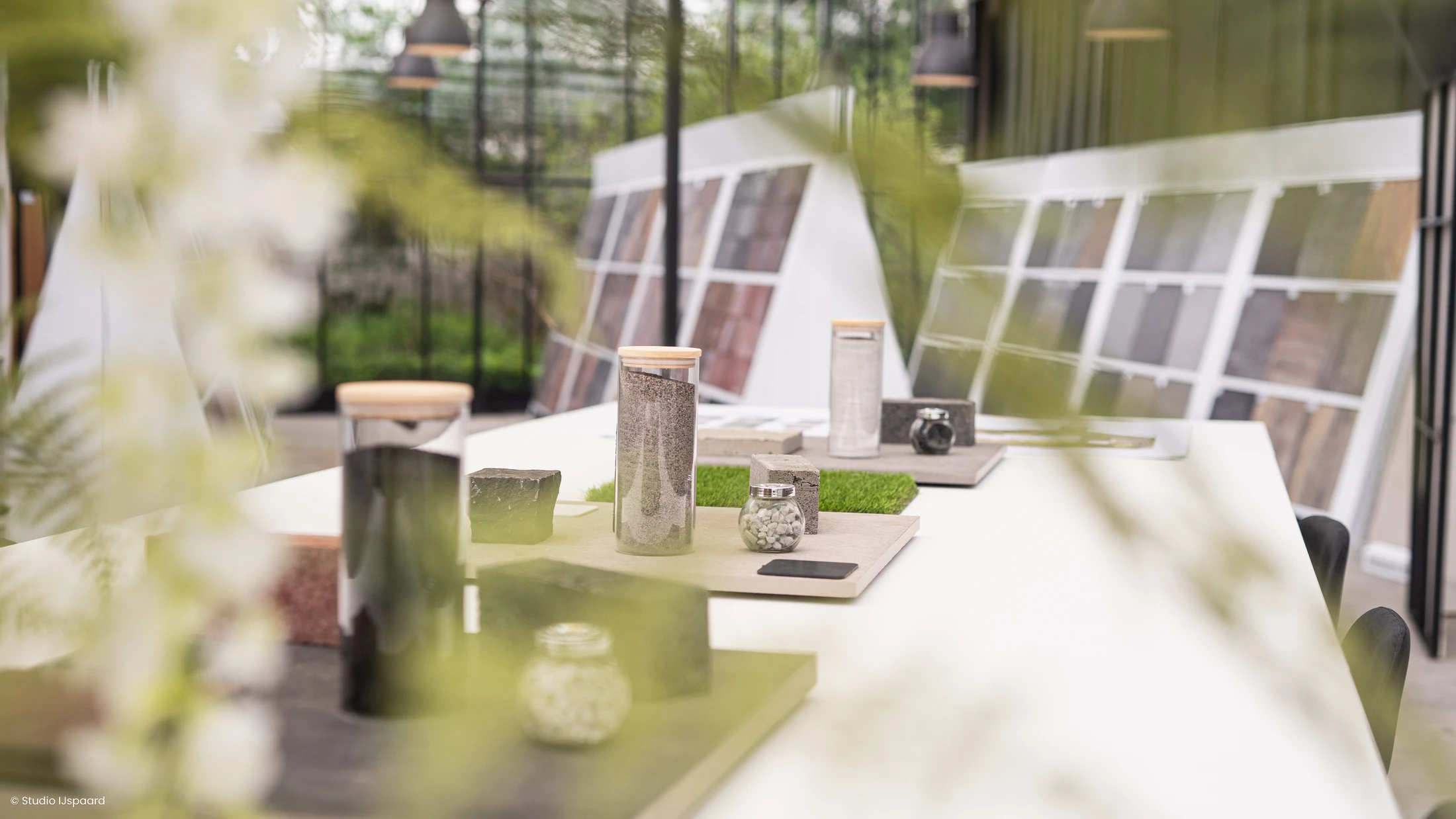 Materiaalmonsters en glazen potten op een lange witte tafel, met presentatierekken op de achtergrond.