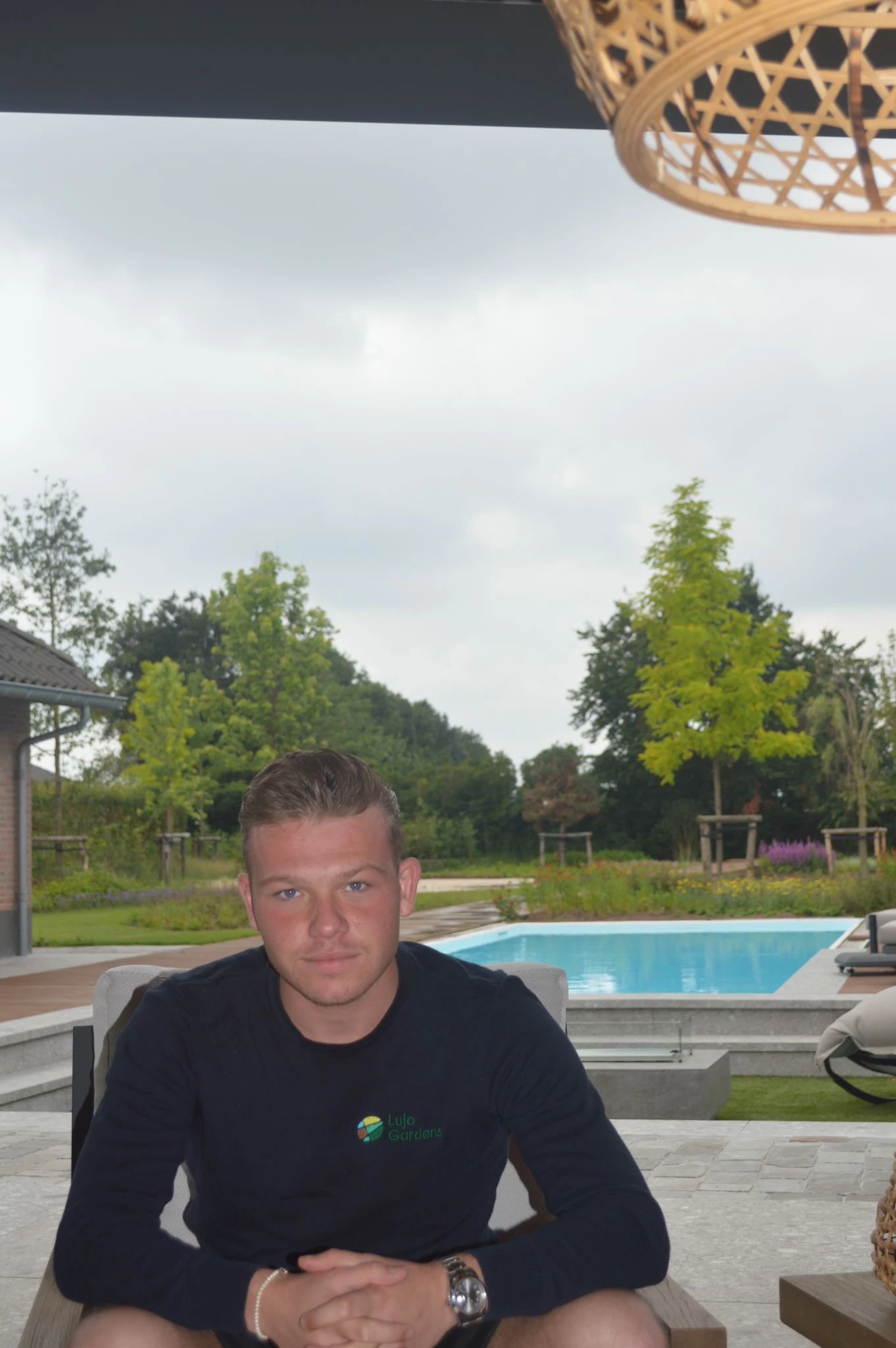 Eigenaar van Lujo Gardens B.V. in een trui met het bedrijfslogo, zittend bij een tuin en zwembad.